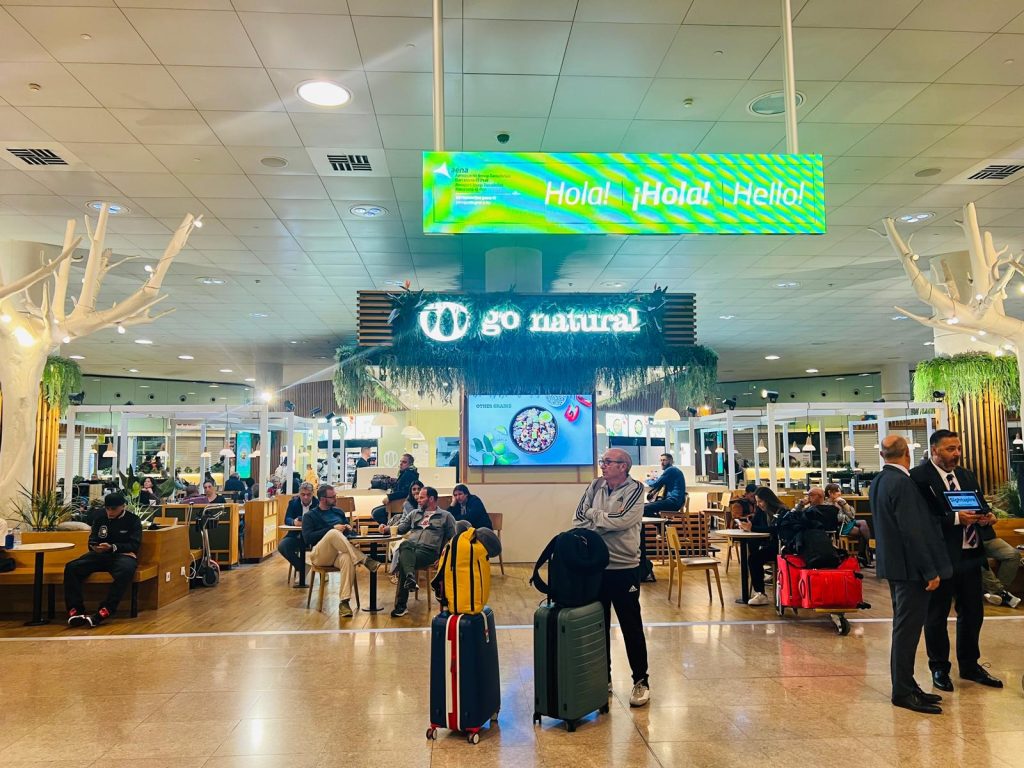 Interior view of Barcelona Airport Terminal 1 showing travelers with suitcases waiting near the “Go Natural” café and a digital sign greeting “Hola! Hello!
